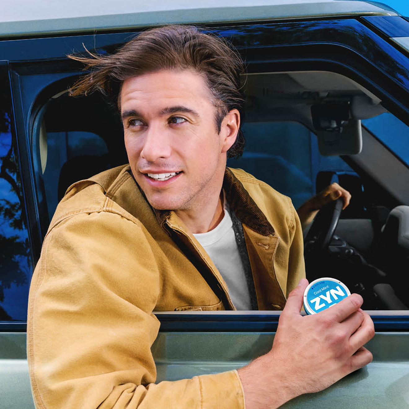 A man leans out of a car window with his head and hand outside, holding a pack of ZYN.