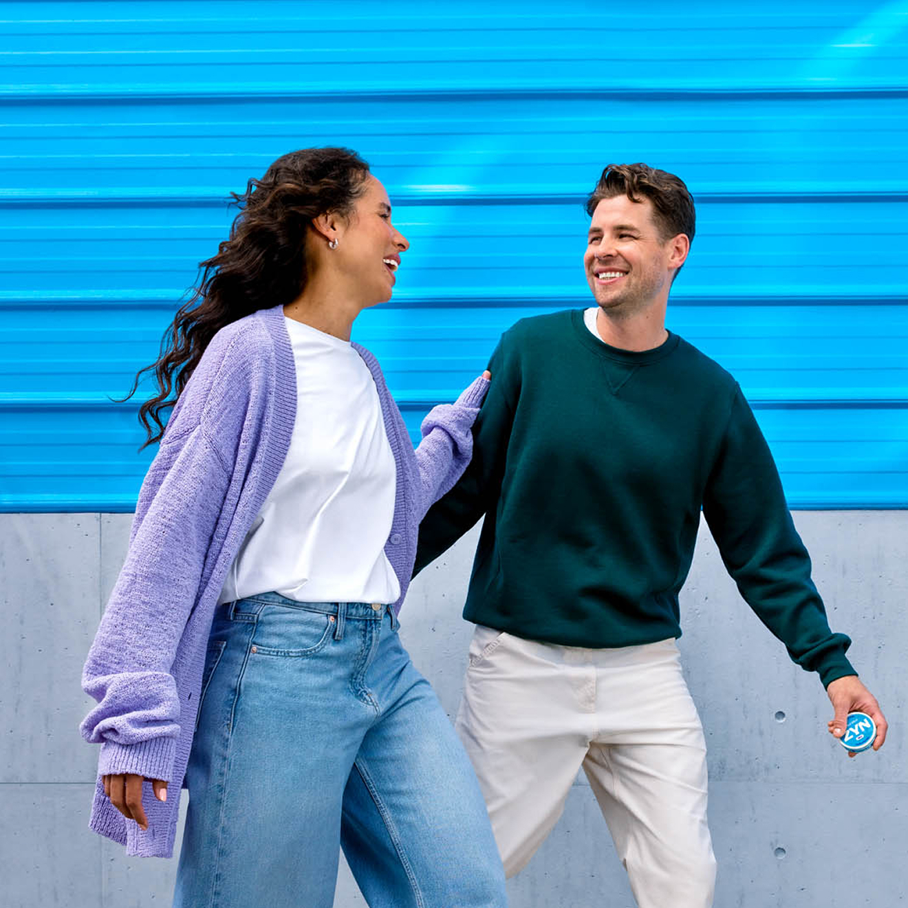 A man and a woman walk down the street, gazing at each other, while the man holds a pack of ZYN.