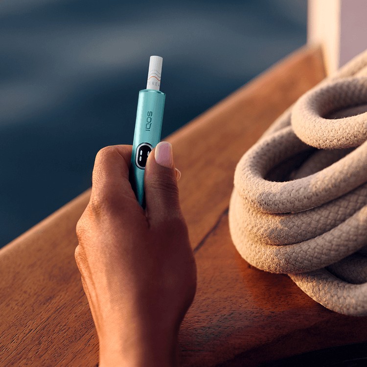 A hand holding an IQOS device with the sea in the background.