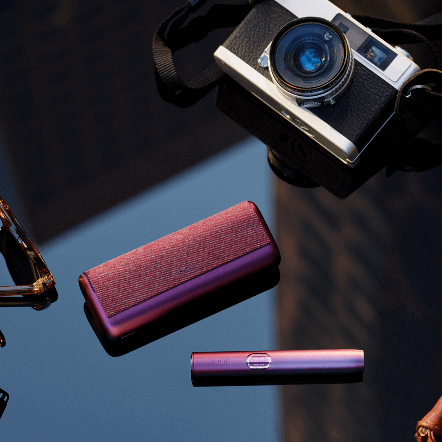 An IQOS ILUMA i PRIME device next to a camera on a glass surface.