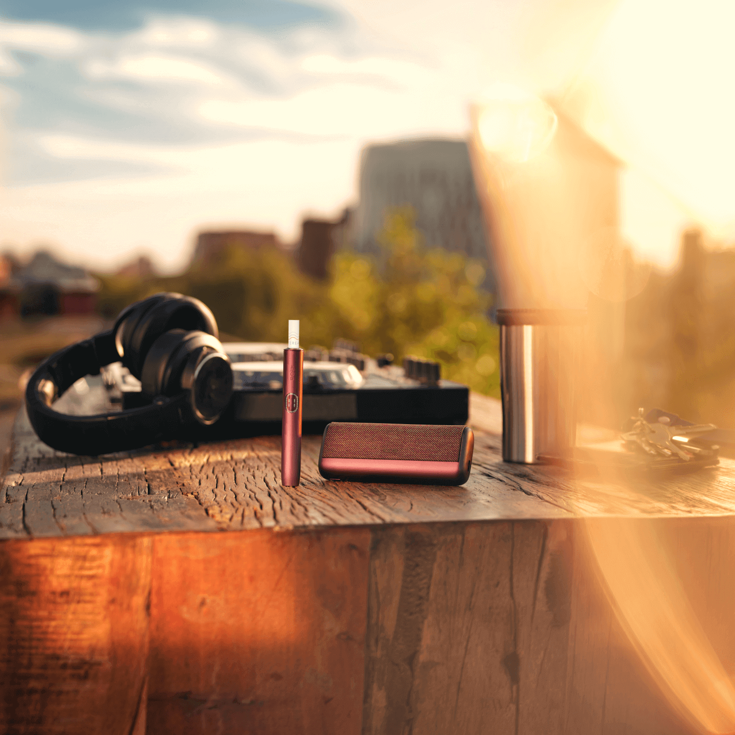A pair of headphones, a portable speaker, a travel mug, a keyring, and an IQOS ILUMA i PRIME device on a wooden surface outdoors in sunlight.