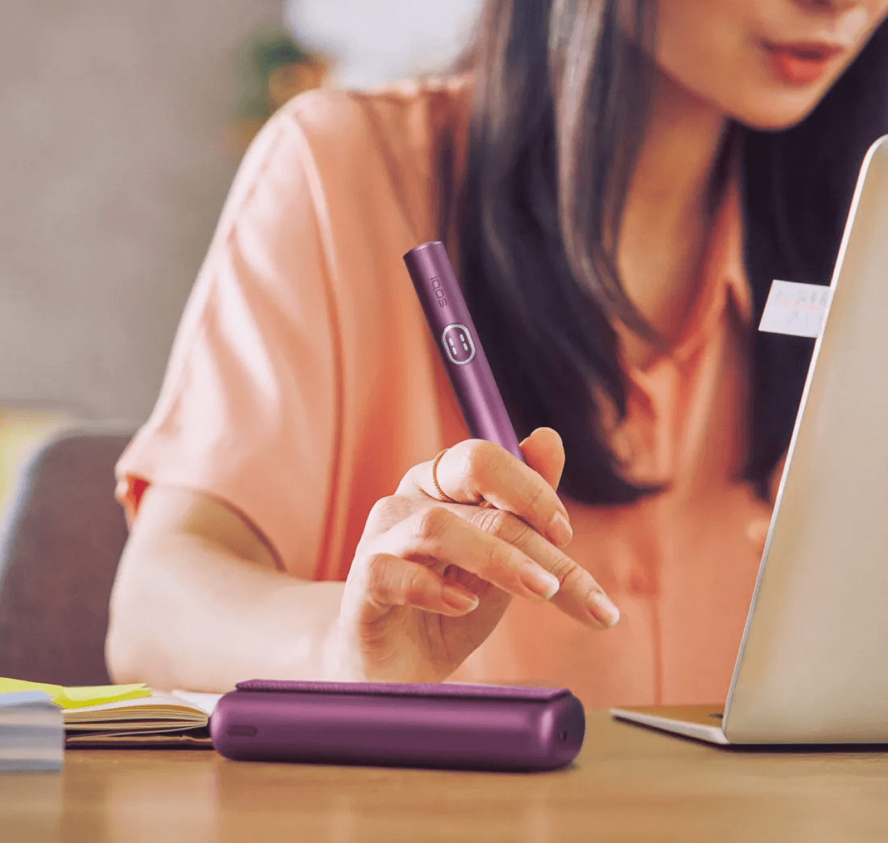 Person working on a laptop and holding a garnet red IQOS ILUMA i PRIME device.