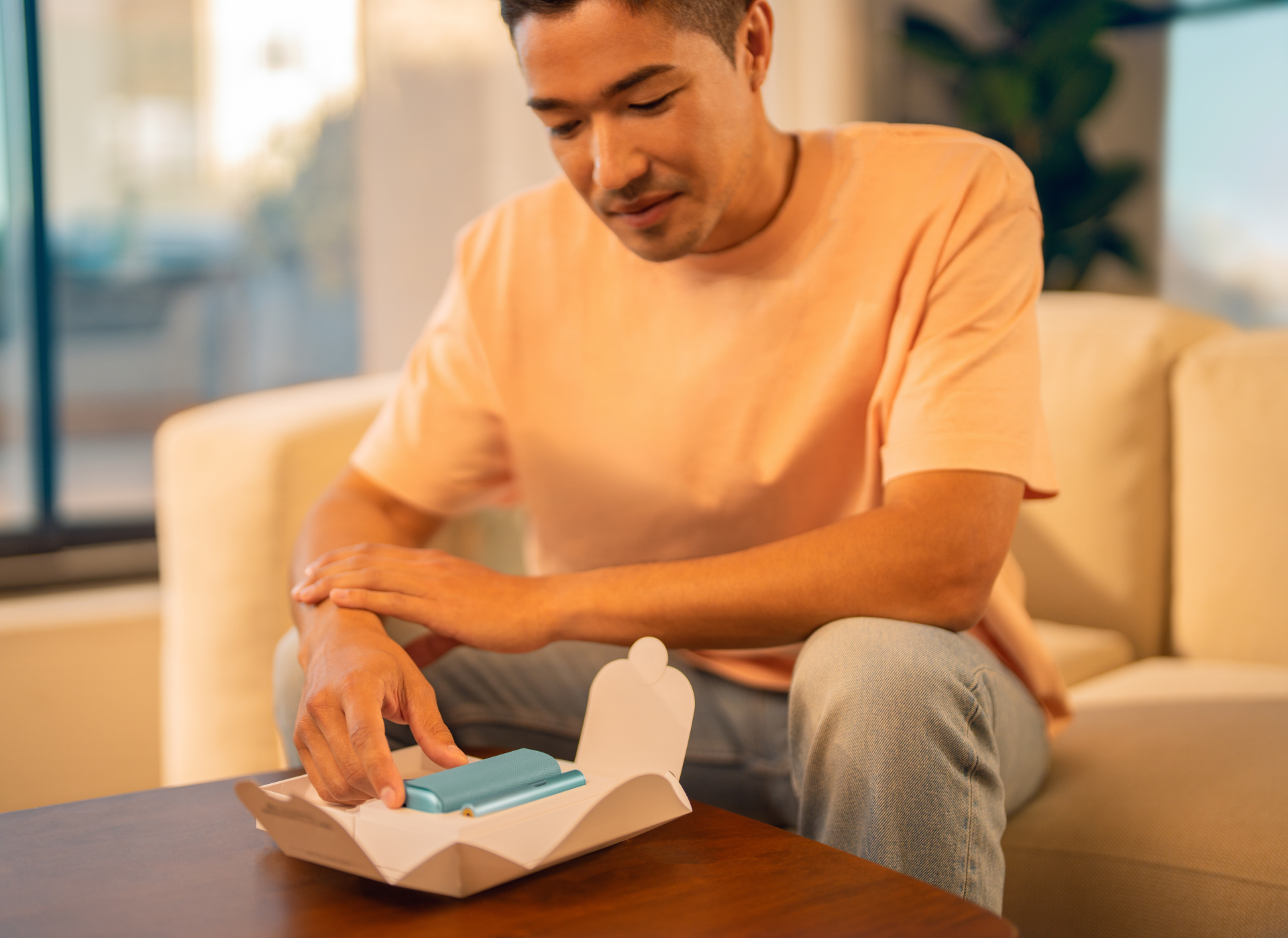 A man is sitting on a couch, unboxing a new IQOS ILUMA i PRIME in Breeze Blue color.