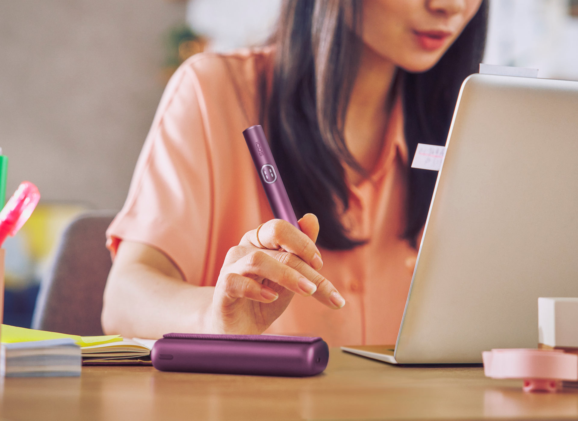 A woman  in front of a laptop, holding an IQOS ILUMA i PRIME holder in Digital Violet color near the Pocket Charger.
