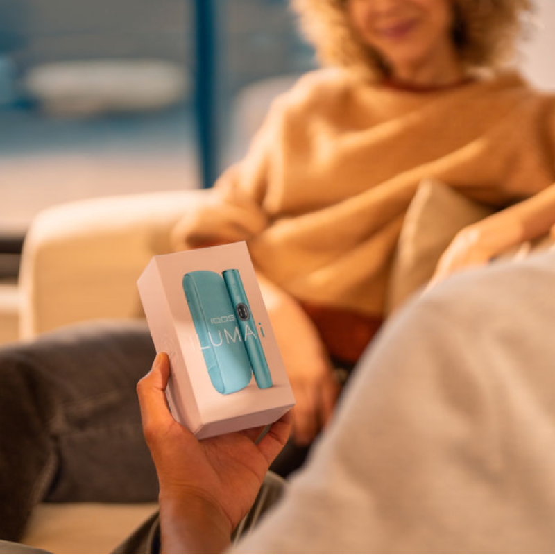 Details of hands holding an IQOS ILUMA i device in front of a laptop.