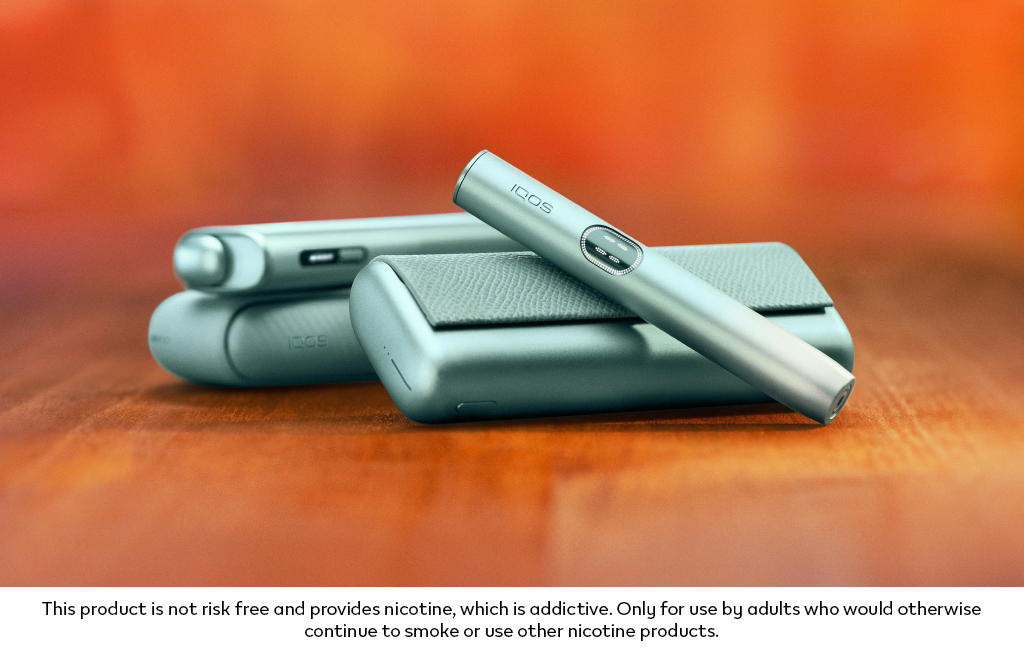 Green IQOS ILUMA i PRIME device above its charger, with an IQOS ILUMA device in the background.