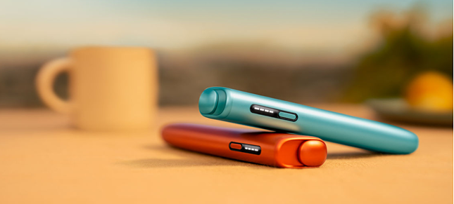 Two IQOS ILUMA i ONE devices. One Terracotta Red and the other Breeze Blue