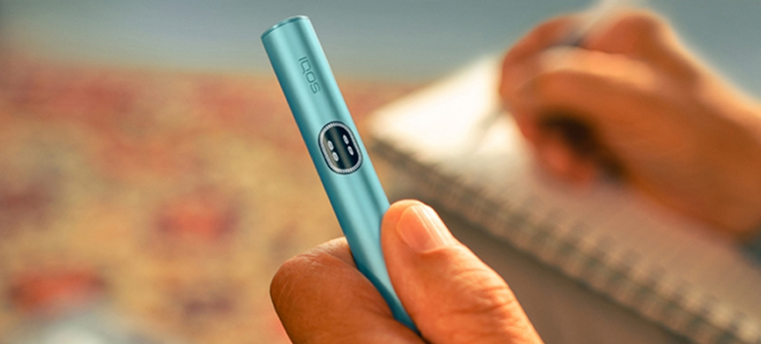Συσκευή IQOS ILUMA i σε χρώμα Breeze Blue, ενώ στο background φαίνεται ένα χέρι να γράφει σε τετράδιο.