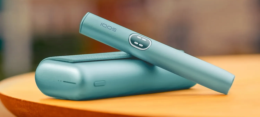 Συσκευή IQOS ILUMA i PRIME με τον φορτιστή της σε χρώμα Breeze Blue, πάνω σε ένα τραπέζι.