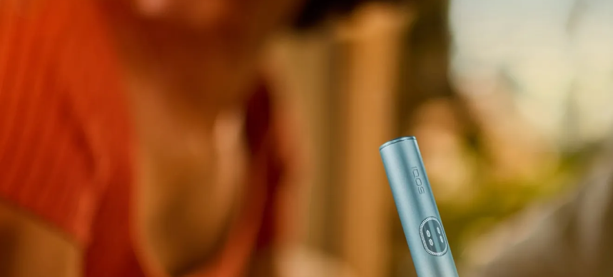Hand holding a Breeze Blue IQOS ILUMA i PRIME device and behind it there is a woman.