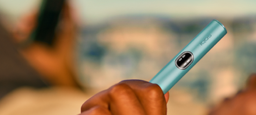 Person lying down, holding a Breeze Blue IQOS ILUMA i device on the left hand and a phone on the right hand.