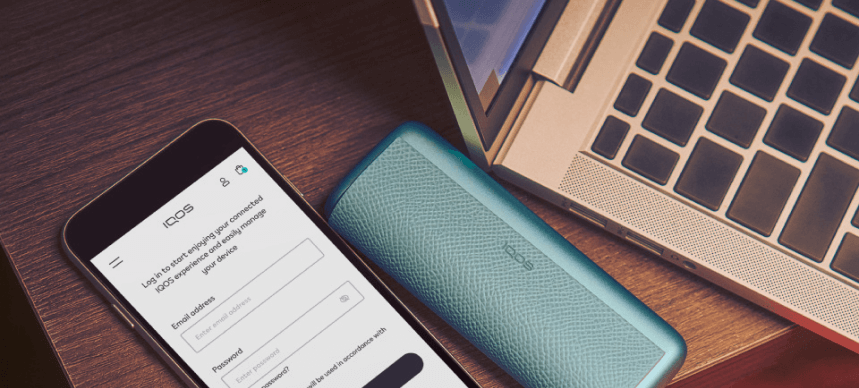 Ένα smartphone που εμφανίζει την οθόνη σύνδεσης στο IQOS, τοποθετημένο πάνω σε ξύλινη επιφάνεια δίπλα σε μια IOQS ILUME i PRIME και ένα ανοιχτό laptop.