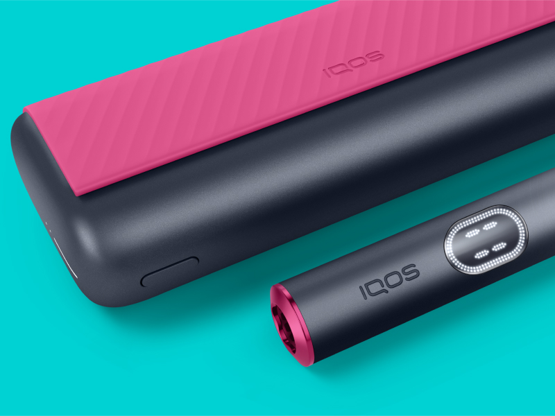 Red and black IQOS ILUMA i PRIME device and its charger on a light turquoise background