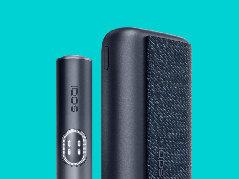 Black IQOS ILUMA i PRIME device and its charger on a light turquoise background