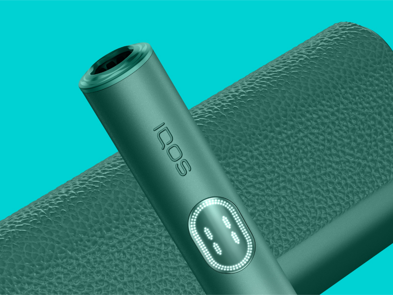 Green IQOS ILUMA i PRIME device and its charger on a light turquoise background