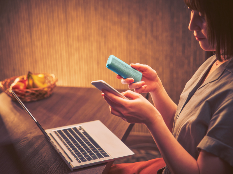 Woman sitting in front of her laptop with a blue IQOS ILUMA i PRIME device and a phone in her hands