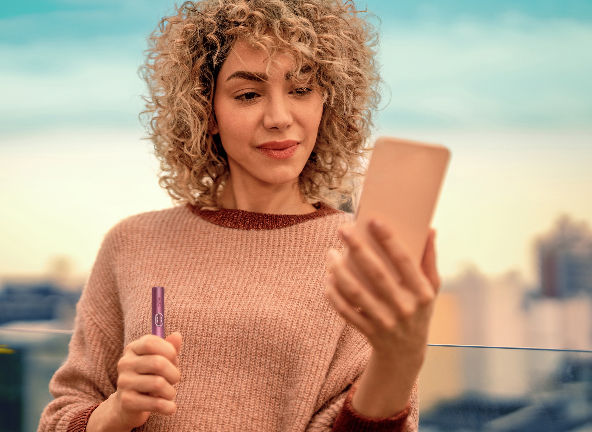 Woman looking at her phone while holding an IQOS ILUMA i PRIME device