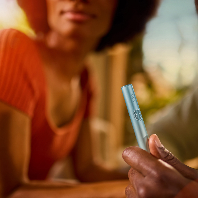 Hand holding an IQOS ILUMA i PRIME device, in front of woman.