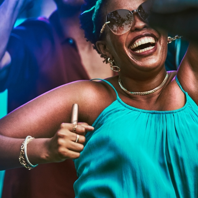 Woman dancing and smiling with a stellar blue IQOS 3 DUO holder in her hand.