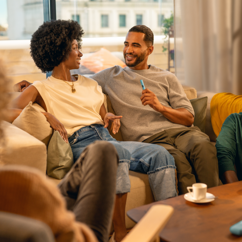 Man holding an IQOS ILUMA i device is sitting on a sofa along with a woman