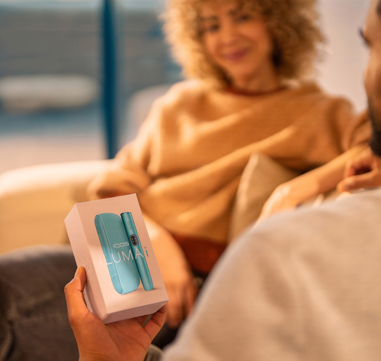 Man holding an IQOS ILUMA i device package, sitting in front of a woman