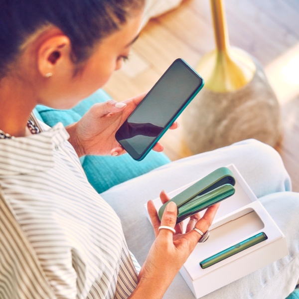 A woman holding a smart phone in her left hand and an IQOS device in her right hand.