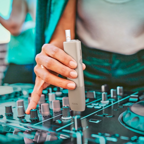 Μία DJ ρυθμίζει τα καντράν σε ένα πικάπ κρατώντας ένα IQOS ILUMA ONE.