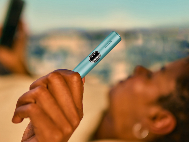 Green IQOS ILUMA i PRIME device and its charger on a light turquoise background