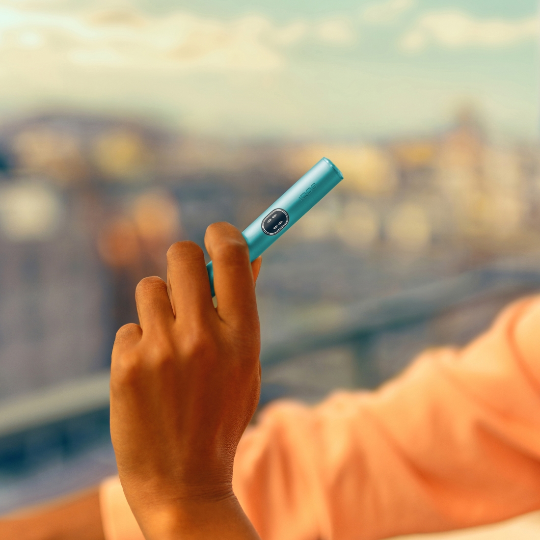 Hand holding an IQOS ILUMA i device, in front of a balcony