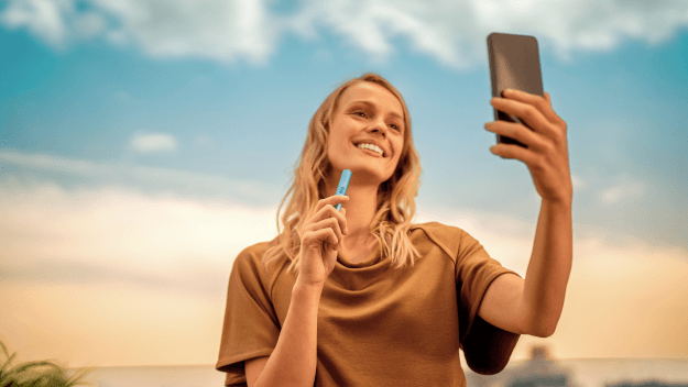 A woman sitting at a table next to her suitcase with a smartphone, passport and plane ticket.