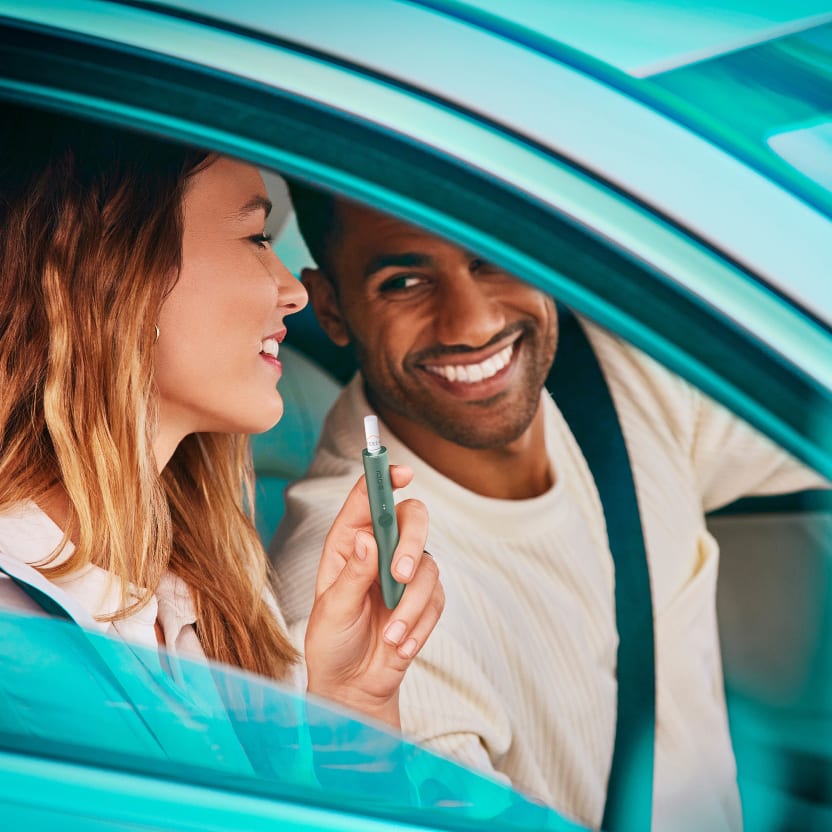 A man and a woman talking in a car. The woman is holding a green IQOS device