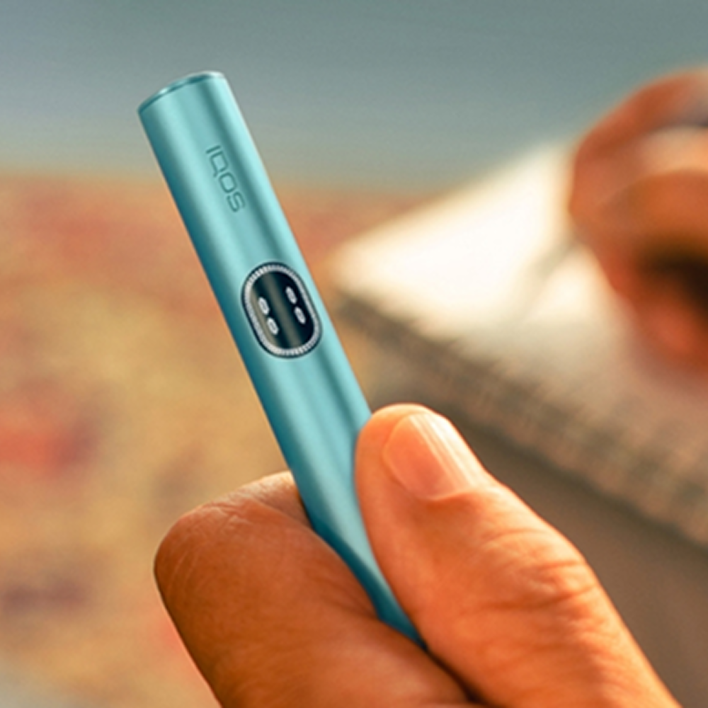 Συσκευή IQOS ILUMA i σε χρώμα Breeze Blue, ενώ στο background φαίνεται ένα χέρι να γράφει σε τετράδιο.