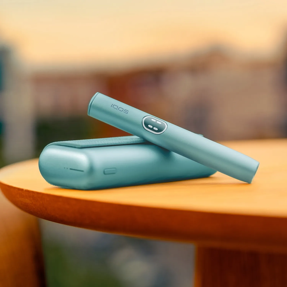 Συσκευή IQOS ILUMA i PRIME με τον φορτιστή της σε χρώμα Breeze Blue, πάνω σε ένα τραπέζι.