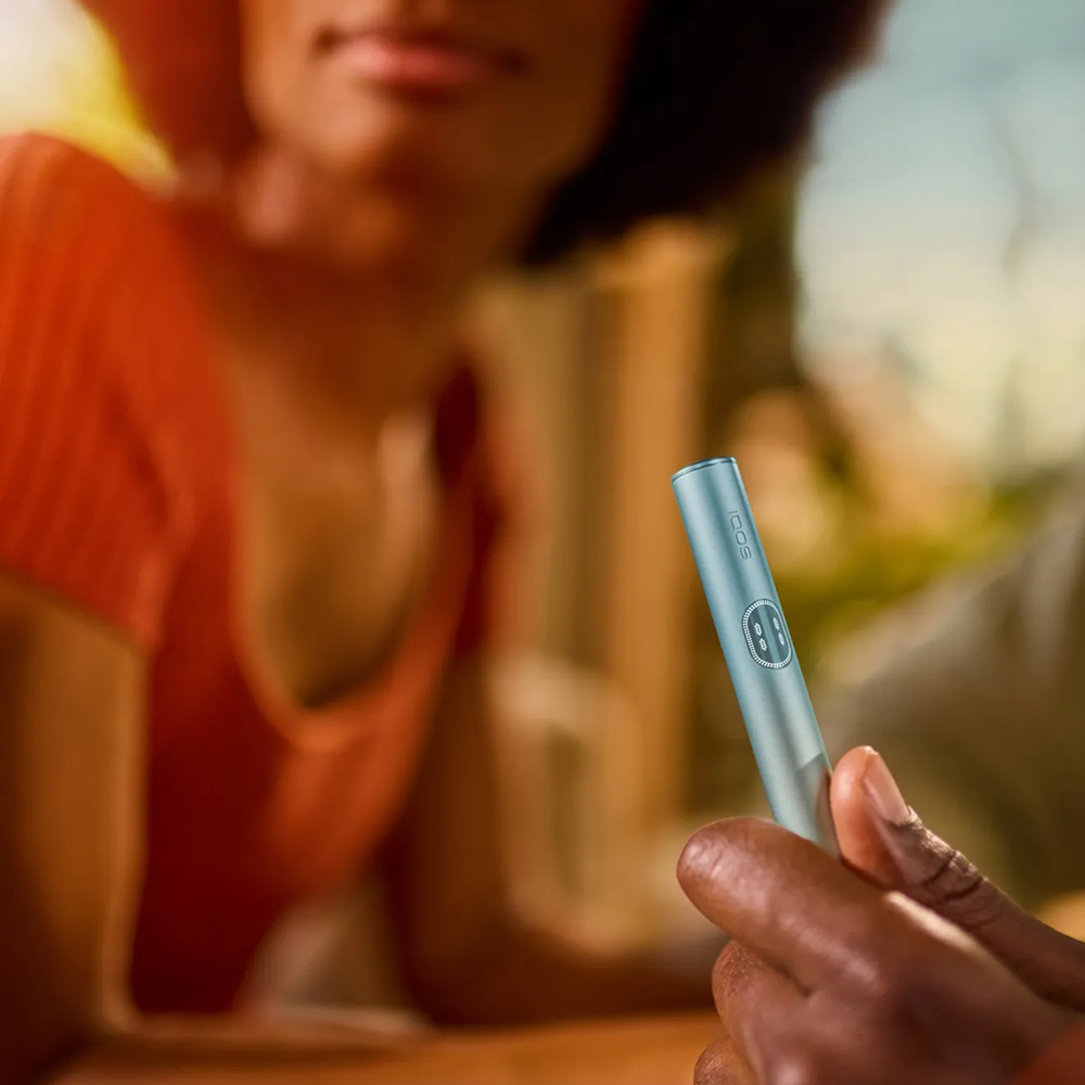 Hand holding a Breeze Blue IQOS ILUMA i PRIME device and behind it there is a woman.