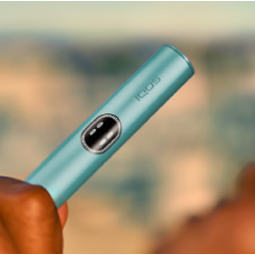 Person lying down, holding a Breeze Blue IQOS ILUMA i device on the left hand and a phone on the right hand.