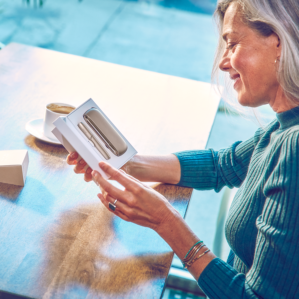 woman opening an IQOS 3 DUO Gold while drinking coffee woman opening an IQOS 3 DUO Gold while drinking coffee