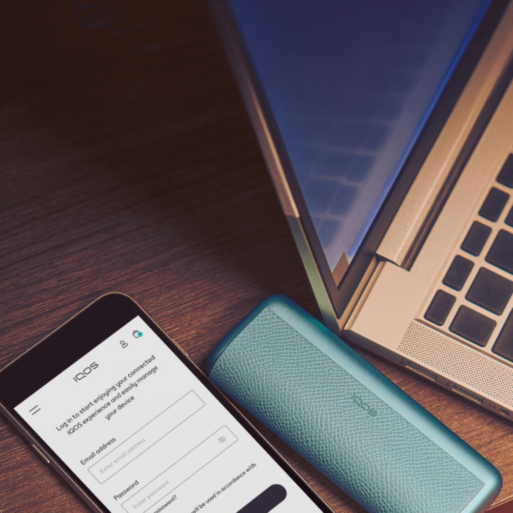 Ένα smartphone που εμφανίζει την οθόνη σύνδεσης στο IQOS, τοποθετημένο πάνω σε ξύλινη επιφάνεια δίπλα σε μια IOQS ILUME i PRIME και ένα ανοιχτό laptop.